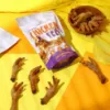 Colorful flat lay of Wiggles N' Nibbles Chicken Feet dog treats 100g pack, surrounded by dried chicken feet and a wooden bowl on a yellow-orange background.